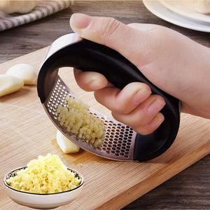 Hand using a garlic press on a wooden cutting board with minced garlic and whole garlic cloves.